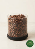 A clear cylindrical container filled with medium roasted coffee beans, sitting on a black circular base against a warm off-white background. A green oval 'COORG INDULGENCE' label is visible in the lower right corner.
