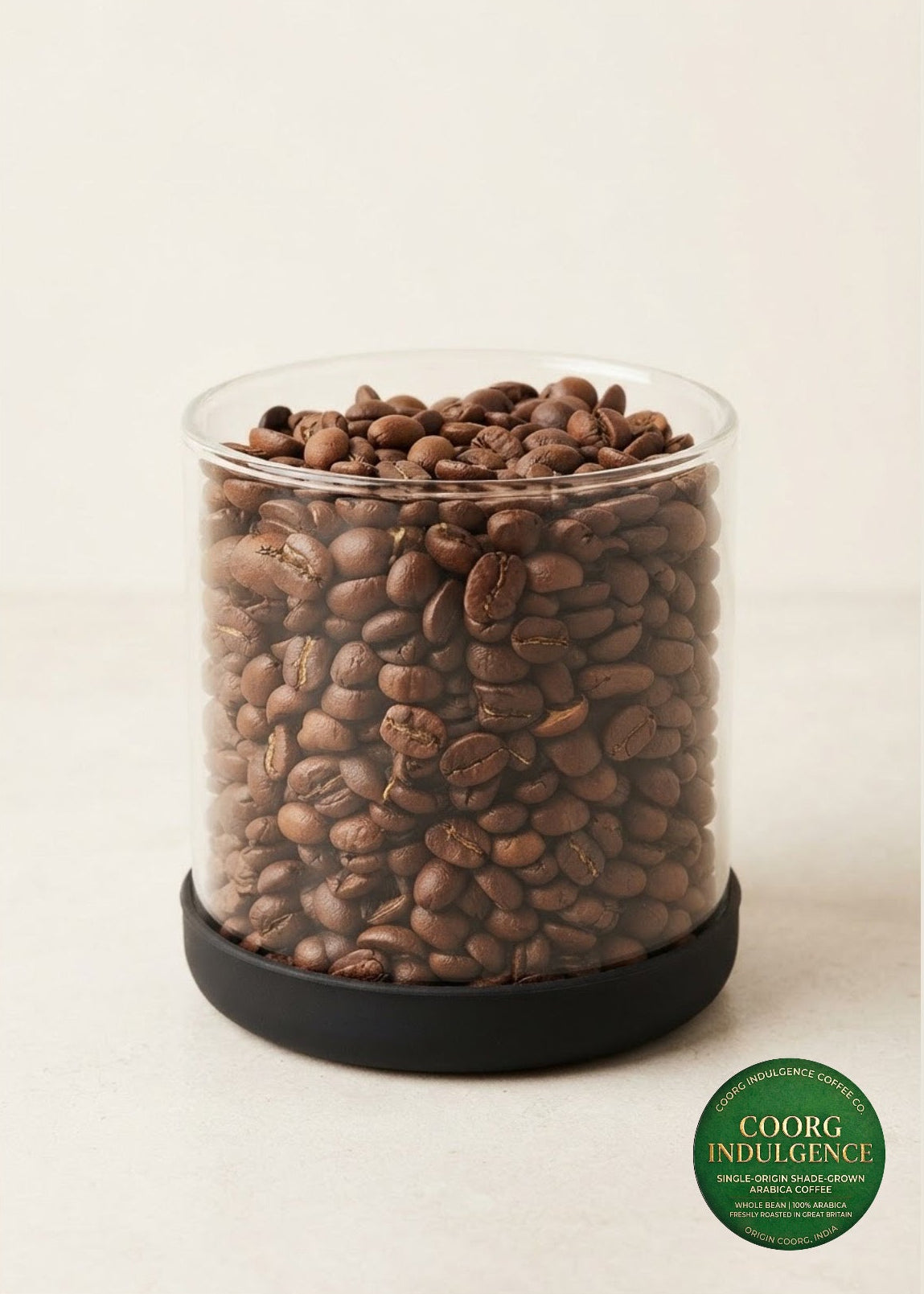 A clear cylindrical container filled with medium roasted coffee beans, sitting on a black circular base against a warm off-white background. A green oval 'COORG INDULGENCE' label is visible in the lower right corner.