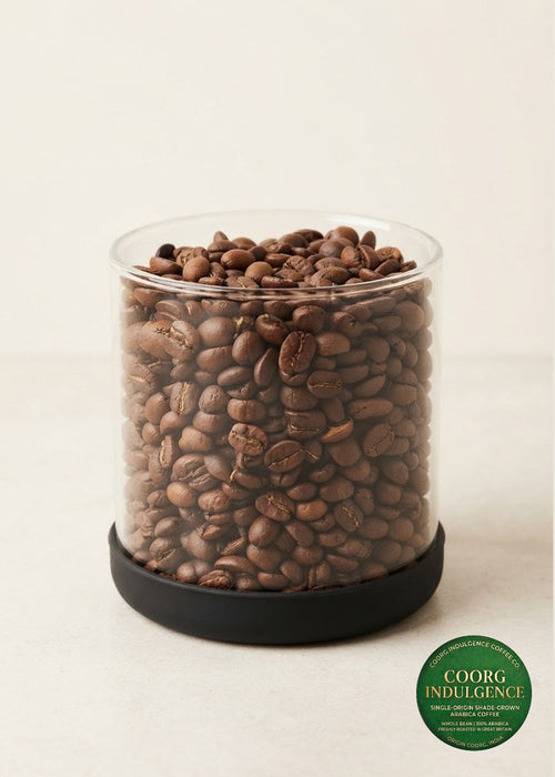 A clear cylindrical container filled with medium roasted coffee beans, sitting on a black circular base against a warm off-white background. A green oval 'COORG INDULGENCE' label is visible in the lower right corner.